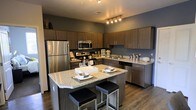 This single-floor space showcases a thoughtfully designed kitchen as its centerpiece. The kitchen features rich, dark wood cabinetry that extends to the upper walls, providing generous storage throughout. Stainless steel appliances, including a full-size refrigerator, microwave, and range, lend a sleek, contemporary look. The countertops are finished in light granite, offering a striking contrast against the darker cabinetry and deep gray walls.

A central kitchen island with matching granite surfaces serves as both a functional workspace and a casual dining area, accommodated by modern black cushioned bar stools. Track lighting above the island highlights the space effectively, complementing the warm wood-toned flooring that runs throughout.

The kitchen layout is designed for efficiency, with ample counter space along the perimeter and a second sink area visible along the side wall. The dark, muted wall tones paired with the warm wood and metallic finishes create a modern yet inviting ambiance, making the kitchen both a practical and visually compelling feature of this space.
