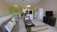 This two-floor residence offers a thoughtfully designed open-concept layout that seamlessly connects the main living spaces. The living room is spacious and inviting, featuring plush carpeting, vaulted ceilings that lend an airy sense of height, and a warm, neutral color palette accented by a bold green feature wall that adds a vibrant pop of color. The living area comfortably accommodates a large sectional sofa, an accent chair with ottoman, and a central coffee table, creating an ideal space for both relaxation and entertaining. Adjacent to the living room, the dining area provides a dedicated space for meals, furnished with a classic table and chairs beneath tasteful wall décor. The kitchen is modern and functional, featuring white cabinetry, a dark granite-style countertop, a breakfast bar with seating, and a full suite of black appliances including a refrigerator and microwave. A hallway off the main living area leads to additional rooms, suggesting a well-organized floor plan with clearly defined private and communal zones. Flush-mount ceiling lighting throughout ensures the space remains bright and well-illuminated. The overall ambiance is clean, contemporary, and livable, with a consistent green and neutral color scheme tying the entire main level together cohesively.