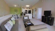 This two-floor residence offers a thoughtfully designed open-concept layout that seamlessly connects the main living spaces. The living room is spacious and inviting, featuring plush carpeting, vaulted ceilings that lend an airy sense of height, and a warm, neutral color palette accented by a bold green feature wall that adds a vibrant pop of color. The living area comfortably accommodates a large sectional sofa, an accent chair with ottoman, and a central coffee table, creating an ideal space for both relaxation and entertaining. Adjacent to the living room, the dining area provides a dedicated space for meals, furnished with a classic table and chairs beneath tasteful wall décor. The kitchen is modern and functional, featuring white cabinetry, a dark granite-style countertop, a breakfast bar with seating, and a full suite of black appliances including a refrigerator and microwave. A hallway off the main living area leads to additional rooms, suggesting a well-organized floor plan with clearly defined private and communal zones. Flush-mount ceiling lighting throughout ensures the space remains bright and well-illuminated. The overall ambiance is clean, contemporary, and livable, with a consistent green and neutral color scheme tying the entire main level together cohesively.