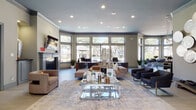 This well-appointed single-story clubhouse offers a sophisticated and welcoming atmosphere designed for both relaxation and productivity. The main lounge serves as the heart of the space, featuring a classic fireplace, a wall-mounted television, and an expansive arrangement of plush seating centered around a sleek glass coffee table atop a large area rug. Recessed lighting illuminates the room, while a bank of wide windows along the rear wall floods the interior with natural light and frames views of the surrounding tree-lined grounds.

Adjacent to the main lounge is a bright café-style seating area enclosed by floor-to-ceiling arched windows, offering sweeping views of an outdoor pool, a covered gazebo, and lush landscaping. This sun-drenched room features light tile flooring, glass-top bistro tables with contemporary chrome-accented chairs, a ceiling fan, and an abundance of potted greenery that enhances the airy, garden-like ambiance.

A dedicated business center provides a quiet and functional workspace, equipped with individual computer workstations and accented by bold blue abstract artwork. The entryway and corridors are finished with light wood-look tile flooring, decorative wall sculptures, and curated artwork, creating a cohesive modern aesthetic that flows throughout the entire space.