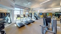 This spacious fitness center is thoughtfully designed to support a comprehensive workout experience. The space is defined by its exposed concrete ceiling, which lends an industrial yet polished character, complemented by sleek linear pendant lighting and herringbone-patterned flooring throughout. Large floor-to-ceiling windows along one wall flood the interior with natural light and provide open views of the surrounding cityscape, creating an energizing atmosphere for exercise. The cardio zone is well-appointed with a row of treadmills, stair climbers, ellipticals, and stationary bikes, many positioned to take full advantage of the exterior views. Multiple flat-screen televisions are mounted throughout the space, keeping members engaged during their workouts. The strength training area offers a diverse selection of equipment, including cable machines, a multi-functional rig, selectorized strength machines, adjustable benches, and free weights neatly organized on dedicated racks. A functional training zone features battle ropes, kettlebells, a punching bag, and a sled, catering to dynamic and high-intensity training styles. Full-length mirrors line portions of the walls, visually expanding the space and supporting proper form. Rolled towels are conveniently stocked near the free weight area, adding a refined, amenity-level touch to the overall facility.