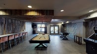 Located at 2121 Delgany St in Denver, Colorado, this amenity space at The Metro Urban Apartments is a thoughtfully designed entertainment and social lounge. The single-floor game room features large-format tile flooring throughout, creating a cohesive and modern foundation that anchors the entire space.

Reclaimed wood wall panels serve as a defining architectural element, lending a warm, industrial character that contrasts beautifully with the textured gray tile accent walls. A statement geometric pendant light hangs above a round high-top table surrounded by cognac leather bar chairs, providing an intimate seating area beneath a wall-mounted flat-screen television. Colorful tile artwork adds a vibrant pop to the neutral palette.

The recreation area is well-equipped with a full-size billiards table as its centerpiece, alongside a foosball table and an arcade game cabinet, offering a variety of entertainment options. A long bar-height counter backed by reclaimed wood paneling provides additional seating along the perimeter, ideal for socializing.

Natural light filters in through windows and a set of glass double doors branded with The Metro logo, which open directly onto an outdoor patio space with additional seating. Recessed ceiling lighting ensures the interior remains well-lit throughout. The overall ambiance blends urban-industrial design with relaxed, functional comfort.