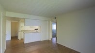 This single-floor space presents an open and functional layout, seamlessly connecting the main living area to the kitchen and a separate room beyond. The living area features wood-style flooring that extends throughout, lending warmth and visual continuity to the space. A textured popcorn ceiling adds dimension overhead, while neutral, light-toned walls create a clean, understated backdrop throughout.

The kitchen is integrated into the space via a partial wall, offering a sense of separation without fully enclosing the area. It features white upper and lower cabinetry, a countertop with a light stone-style finish, a sink, and a range, providing a practical and organized cooking environment. Under-cabinet lighting adds subtle illumination to the workspace.

A hallway visible from the main living area leads to an additional room, which appears to be carpeted and benefits from natural light entering through a window. A ceiling vent is also visible, indicating a central HVAC system. Overall, the space offers a straightforward, well-maintained interior with a bright, neutral aesthetic suited to a variety of uses.
