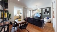 This well-appointed single-floor residence combines modern design with functional living across a thoughtfully arranged open-concept layout. Warm hardwood flooring flows throughout, tying together the living, dining, and kitchen areas seamlessly.

The living room serves as the focal point of the space, anchored by a deep navy tufted sofa and accented by a striking mid-century modern chandelier with brass detailing. A dark accent wall provides a bold backdrop for curated wall art and a large flat-screen TV. A glass-paneled door opens directly to a private balcony, filling the room with natural light and offering views of the surrounding urban environment.

Adjacent to the living area, the dining space accommodates a four-person wood-top table, with direct sightlines into the kitchen. The kitchen features light wood cabinetry, stainless steel appliances including a refrigerator, range, and microwave, and a clean white tile backsplash.

A built-in nook with open shelving and a dedicated desk area provides a compact yet functional workspace tucked neatly off the main living area.

The bathroom is finished with granite countertops, a single undermount sink set in light wood vanity cabinetry, and a full soaking tub with a tiled surround and a curved shower rod. Neutral tile flooring and a framed mirror complete the polished look.
