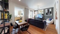 This well-appointed single-floor residence combines modern design with functional living across a thoughtfully arranged open-concept layout. Warm hardwood flooring flows throughout, tying together the living, dining, and kitchen areas seamlessly.

The living room serves as the focal point of the space, anchored by a deep navy tufted sofa and accented by a striking mid-century modern chandelier with brass detailing. A dark accent wall provides a bold backdrop for curated wall art and a large flat-screen TV. A glass-paneled door opens directly to a private balcony, filling the room with natural light and offering views of the surrounding urban environment.

Adjacent to the living area, the dining space accommodates a four-person wood-top table, with direct sightlines into the kitchen. The kitchen features light wood cabinetry, stainless steel appliances including a refrigerator, range, and microwave, and a clean white tile backsplash.

A built-in nook with open shelving and a dedicated desk area provides a compact yet functional workspace tucked neatly off the main living area.

The bathroom is finished with granite countertops, a single undermount sink set in light wood vanity cabinetry, and a full soaking tub with a tiled surround and a curved shower rod. Neutral tile flooring and a framed mirror complete the polished look.