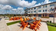 Located at 17326 N Eldridge Pkwy in Tomball, Texas, this well-appointed outdoor courtyard is designed to foster community and comfortable social interaction. The space is organized around two rectangular fire pit tables, each surrounded by bold orange Adirondack chairs that create a striking, welcoming atmosphere. Strips of lush artificial turf run between the seating areas, providing a clean visual contrast against the light-toned paver flooring that extends throughout the courtyard. Overhead, festoon string lights are strung across the area, adding a warm, inviting ambiance suitable for both daytime and evening gatherings. The courtyard is enclosed by decorative wrought iron fencing, offering a sense of privacy while maintaining open sightlines to the surrounding community. A white board-and-batten building anchors one end of the space, featuring double doors and brick accents at its base. Mature trees and manicured landscaping line the perimeter, softening the hardscape elements. A covered pavilion structure is visible toward the rear of the property, extending the outdoor amenity offerings. The overall design balances functionality with a lively aesthetic, making it a versatile destination for casual relaxation or organized outdoor events.