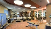 This fitness center is a thoughtfully designed exercise facility that combines functionality with a polished, upscale aesthetic. The space is defined by its striking dropped ceiling, finished in a rich dark tone and adorned with large, circular flush-mount light fixtures that cast a bright, even glow across the entire floor. The warm-hued, patterned carpet underfoot adds a layer of visual interest while complementing the sophisticated color palette of the room.

Floor-to-ceiling mirrors line the walls, effectively expanding the sense of space and allowing users to monitor their form throughout workouts. Structural columns are seamlessly integrated into the layout, adding to the architectural character of the room.

The facility is outfitted with an extensive selection of cardio equipment, including treadmills, elliptical trainers, and stationary bikes, all arranged in well-organized rows. A dedicated free weights area with benches and a full dumbbell rack provides ample options for strength training. Additional resistance and cross-training machines are positioned throughout the space, ensuring a complete workout experience.

The overall ambiance is energetic yet refined, with the combination of modern lighting, mirrored surfaces, and a carefully curated equipment layout creating an inviting and motivating environment for users of all fitness levels.