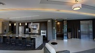 This sophisticated single-floor space showcases a modern open-concept design that seamlessly integrates the kitchen and living areas. The kitchen is a standout feature, equipped with dark cabinetry, stainless steel appliances including a double wall oven and refrigerator, and a generous island topped with striking veined stone countertops. Pendant lighting and warm recessed cove lighting along the ceiling add depth and ambiance to the space. The ceiling itself features architectural tray detailing with integrated accent lighting, contributing to the refined atmosphere.

Bar-style seating lines the kitchen island, making it ideal for casual dining or entertaining. Adjacent to the kitchen, the living area includes contemporary upholstered seating and a minimalist round side table, maintaining the clean aesthetic throughout.

Large glass doors at the rear open toward a bright exterior space, allowing natural light to filter into the interior and providing a visual connection to the outdoors. Decorative artwork and subtle architectural wall treatments add character to the hallway and connecting spaces. The overall ambiance is polished and contemporary, with a cohesive palette of dark wood tones, neutral grays, and metallic accents.