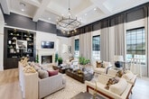 Located at 14086 Airline Hwy in Gonzales, Louisiana, this single-floor space makes a striking architectural statement through its refined design and thoughtful layout. The main lounge area commands attention with soaring coffered ceilings accented in deep charcoal, anchored by a dramatic wrought-iron chandelier that serves as a centerpiece. Stately arched columns and detailed crown molding frame the open floor plan, while light hardwood flooring runs seamlessly throughout, lending warmth and continuity to the space. Floor-to-ceiling drapery panels flank large multi-pane windows, flooding the interior with natural light and offering views to the exterior. A classic fireplace with a white surround adds a focal point to one wall, complemented by curated wall art and gold-toned accent fixtures. The lounge transitions naturally into a functional office area, which features a large glass viewing window into a well-stocked interior room, a reception-style console, and a private workspace with a dark wood desk and modern seating. Arched doorways connect the areas fluidly, maintaining the space's cohesive, upscale aesthetic. The overall ambiance is sophisticated and polished, blending classical architectural details with contemporary interior design for a truly distinguished environment.