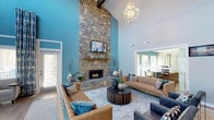 This thoughtfully designed single-floor space makes an immediate impression with its soaring double-height ceilings and bold design choices. The centerpiece of the main lounge area is a striking floor-to-ceiling stacked stone fireplace, anchoring the room with a sense of grandeur and warmth. A rustic wooden mantel and hearth ledge add a natural, grounded contrast to the dramatic vertical scale of the stonework, while a mounted flat-screen television above the mantel blends modern convenience with classic design.

The walls are dressed in a vivid teal blue, creating an energetic yet inviting atmosphere that is complemented by exposed dark ceiling beams overhead. An elegant crystal chandelier hangs above, casting ambient light throughout the space. The seating area is anchored by a large area rug and features tufted leather sofas accented with teal and neutral throw pillows, arranged around a substantial dark wood round coffee table — a perfect setting for relaxed gatherings.

Natural light filters in through multiple windows and glass-paneled openings, connecting the main lounge to an adjacent room that appears to serve as a dining or meeting area. The overall ambiance is one of upscale comfort, blending rustic textures with contemporary style and a cohesive, vibrant color palette.