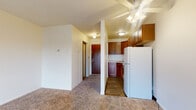 This single-floor unit offers a practical and functional layout designed to maximize usable space. The living area is generously proportioned, featuring neutral carpet flooring, a ceiling fan with integrated lighting, and a baseboard heating unit along the wall. A sliding glass door floods the room with natural light and provides direct access to a private balcony with outdoor views of surrounding greenery.

The kitchen is positioned adjacent to the living space, partially separated by a structural column that defines the two areas. It features warm-toned wood cabinetry both above and below the counters, a light-colored countertop, a stainless steel sink, an electric coil range with overhead ventilation hood, and a full-size white refrigerator. The kitchen floor is finished with wood-look vinyl planking, contrasting with the carpet in the living area.

The bathroom includes a wood-cabinet vanity with a clean white countertop and integrated sink, chrome fixtures, a large mirror, and a tiled shower and tub combination. The entry hallway connects the front door to the main living space and kitchen, providing a clear and organized flow throughout the unit.