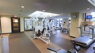 This fitness center offers a thorough and well-organized workout environment designed to accommodate a wide range of exercise needs. The space is anchored by a robust selection of strength training equipment, including multi-station cable machines and a variety of resistance and weight machines arranged to allow comfortable movement between stations. Full-length mirrors line one wall, enhancing the sense of openness and allowing users to monitor their form during workouts. The flooring transitions between cushioned rubber matting in the strength zones and hardwood-style surfaces in the cardio area, providing both safety and functionality. The cardio section is positioned near large windows that invite natural light and offer views to the outdoors, creating an energizing atmosphere. Ellipticals, stationary bikes, and additional cardio machines are neatly arranged in this brighter section. Overhead, a tray ceiling with recessed lighting adds architectural depth and ensures the space is evenly and warmly illuminated throughout. The overall ambiance is clean, modern, and purposeful, reflecting a professional-grade facility that balances practicality with a welcoming design aesthetic suited for daily use.