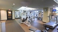 This fitness center offers a thorough and well-organized workout environment designed to accommodate a wide range of exercise needs. The space is anchored by a robust selection of strength training equipment, including multi-station cable machines and a variety of resistance and weight machines arranged to allow comfortable movement between stations. Full-length mirrors line one wall, enhancing the sense of openness and allowing users to monitor their form during workouts. The flooring transitions between cushioned rubber matting in the strength zones and hardwood-style surfaces in the cardio area, providing both safety and functionality. The cardio section is positioned near large windows that invite natural light and offer views to the outdoors, creating an energizing atmosphere. Ellipticals, stationary bikes, and additional cardio machines are neatly arranged in this brighter section. Overhead, a tray ceiling with recessed lighting adds architectural depth and ensures the space is evenly and warmly illuminated throughout. The overall ambiance is clean, modern, and purposeful, reflecting a professional-grade facility that balances practicality with a welcoming design aesthetic suited for daily use.