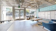 Located at 1 N Fifth Ave in downtown Tucson, Arizona, this residential lobby offers a well-appointed, single-floor open-plan space that blends industrial and contemporary design. The exposed metal mesh ceiling reveals structural systems overhead while recessed and pendant lighting illuminate the polished concrete floors below. A bold teal geometric-patterned accent wall provides a vibrant backdrop to a full bank of resident mailboxes, serving as a functional centerpiece of the lobby. Dual elevators are conveniently positioned nearby for resident access. The seating areas are furnished with stylish mid-century-inspired blue armchairs arranged around a glass-top table, set atop a layered area rug with abstract patterning. A warm-toned slatted wood feature wall anchors a secondary lounge zone, accented by colorful artwork and additional seating options including a bench and counter-height stool. Large glass entry doors face the street, flooding the space with natural light and offering views of the surrounding downtown Tucson streetscape. A luggage cart station and a vending machine round out the practical amenities of this thoughtfully designed, welcoming lobby environment.