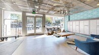 Located at 1 N Fifth Ave in downtown Tucson, Arizona, this residential lobby offers a well-appointed, single-floor open-plan space that blends industrial and contemporary design. The exposed metal mesh ceiling reveals structural systems overhead while recessed and pendant lighting illuminate the polished concrete floors below. A bold teal geometric-patterned accent wall provides a vibrant backdrop to a full bank of resident mailboxes, serving as a functional centerpiece of the lobby. Dual elevators are conveniently positioned nearby for resident access. The seating areas are furnished with stylish mid-century-inspired blue armchairs arranged around a glass-top table, set atop a layered area rug with abstract patterning. A warm-toned slatted wood feature wall anchors a secondary lounge zone, accented by colorful artwork and additional seating options including a bench and counter-height stool. Large glass entry doors face the street, flooding the space with natural light and offering views of the surrounding downtown Tucson streetscape. A luggage cart station and a vending machine round out the practical amenities of this thoughtfully designed, welcoming lobby environment.