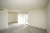 This single-floor residential unit offers a clean, functional layout with a cohesive neutral color palette throughout. The entry corridor welcomes you with plush carpeting underfoot, smooth cream-toned walls, and a wall-mounted intercom panel — a practical feature for everyday use. A solid white front door with double hardware provides a secure entry point, and a ceiling-mounted light fixture keeps the passage well-lit.

The living area is generously proportioned, featuring soft wall-to-wall carpeting and a ceiling fan that promotes comfortable airflow. Structural beams define the space subtly, and an open passage leads directly into the kitchen, which is equipped with a full-size refrigerator and light-toned cabinetry. The layout encourages an easy flow between the living and kitchen areas.

The bedroom is a well-sized, open room with continued carpeting and cream walls, offering a versatile blank canvas. A wall switch and electrical outlets are conveniently placed throughout.

The bathroom is finished with warm beige tile flooring and features a full-size soaking tub with a tiled surround, a shower rod, a toilet, and a white vanity with under-cabinet storage. Overall, the unit presents a straightforward, move-in-ready interior with consistent finishes and practical design elements throughout.
