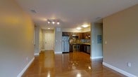 This single-floor space showcases an open-concept layout that seamlessly integrates the living area with the kitchen. The flooring throughout is polished hardwood, reflecting the overhead lighting and contributing to a warm, inviting ambiance.

The kitchen is well-appointed with stainless steel appliances, including a full-size refrigerator, a range with oven, and an over-the-range microwave. Dark-toned wood cabinetry lines the walls, complemented by granite or stone countertops that extend to provide ample workspace. A utility panel is visible on the adjacent wall, and the kitchen benefits from dedicated overhead lighting that illuminates the workspace effectively.

A hallway extends from the main living area, leading to additional rooms. The corridor features built-in storage or closet space along one side and is finished with the same hardwood flooring. A white entry door is visible at the end of the hallway, providing a clear sense of the layout's depth.

The living space features neutral-toned walls, recessed lighting, and a track lighting fixture, ensuring consistent illumination. The overall design is contemporary and functional, with clean lines and practical finishes throughout.