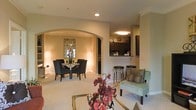 This single-floor residence offers an open-concept layout that seamlessly connects its key living areas, creating a cohesive and welcoming atmosphere throughout. The living area serves as the heart of the space, featuring plush carpeting, recessed lighting, and stylish furnishings that establish a warm, sophisticated ambiance.

A graceful arched entryway draws the eye into the dedicated dining room, where a round dining table surrounded by sleek, upholstered chairs is elegantly framed by soft accent lighting and decorative wall art. The arch adds a refined architectural detail that lends the space a sense of understated elegance.

Adjacent to the living and dining areas, the kitchen is accessible via a pass-through counter with bar stool seating, making it ideal for casual dining and entertaining. The kitchen features dark cabinetry that contrasts beautifully with the lighter tones found throughout the rest of the space. Overhead lighting brightens the workspace, while the open counter connection keeps the cook engaged with guests in the main living area.

Throughout the residence, warm neutral tones on the walls complement the carpeted floors, while a combination of recessed and ambient lighting ensures each area feels inviting. The overall design balances functionality with tasteful décor, resulting in a comfortable and well-appointed living environment.