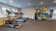 This fitness center offers a comprehensive workout environment designed to accommodate a wide range of exercise needs. The space is finished with durable rubber tile flooring throughout, providing both safety and functionality for various training activities. Recessed lighting illuminates the room evenly, supplemented by a large ceiling fan that promotes air circulation during intense workouts.

Multiple windows allow natural light to fill the space and provide views of the surrounding exterior, creating an open and energizing atmosphere. French doors connect the fitness center to an adjacent area, enhancing the flow between spaces.

The cardio zone features several treadmills and an elliptical trainer, while the strength training area is equipped with a multi-station cable machine, a leg curl/extension machine, and a dedicated free weight section with a full dumbbell rack and adjustable bench. A large wall-mounted mirror expands the sense of space and allows for form monitoring during exercise.

Functional training accessories, including stability balls, resistance bands, and step platforms, are neatly organized on a storage rack beside the mirror. Additional amenities include a wall-mounted television, a water fountain, a hand sanitizer dispenser, and posted fitness center rules, making this a practical and well-appointed facility.