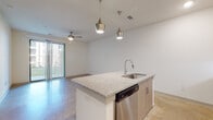 This single-floor unit showcases a contemporary, open-concept design with a neutral color palette and polished concrete floors throughout. The kitchen serves as the centerpiece, featuring a large island topped with granite countertops, an integrated sink, and a stainless steel dishwasher beneath. Pendant lighting with metallic finishes hangs above the island, adding a modern aesthetic. Cabinetry in a warm wood tone lines the kitchen walls, complemented by a full suite of stainless steel appliances including a French door refrigerator, microwave, and range. The living area flows seamlessly from the kitchen and opens to a private balcony through vertical blind-covered sliding glass doors, with a ceiling fan overhead for year-round comfort.

The bathroom is well-appointed with a long wood-toned vanity, granite countertop, and a glass-enclosed walk-in shower. A generously sized walk-in closet features multi-tiered wire shelving for ample storage. The bedroom is a bright, quiet space with a large window allowing natural light. A dedicated laundry room houses a full-size washer and dryer alongside the water heater, rounding out the practical amenities of this well-designed space.