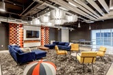 Located at 720 Main St in the heart of Kansas City, this single-floor amenity space delivers a striking blend of industrial architecture and bold, contemporary design. Exposed black ceilings with visible ductwork and conduit run overhead, while polished concrete floors ground the open-plan layout with a clean, urban aesthetic.

The space is organized into several distinct yet connected zones. A lounge area features deep navy blue sofas and vibrant yellow accent chairs arranged around an electric fireplace, with a large-format TV mounted on a bold checkered tile feature wall. Geometric acoustic panels hang from the ceiling, adding visual texture while enhancing the room's acoustics. Floor-to-ceiling glass walls introduce natural light and connect this zone to adjacent areas.

A central gathering zone offers a natural wood dining table surrounded by bright orange chairs, set atop a vivid, wave-patterned area rug. Nearby, a welcoming seating vignette with an orange sofa and neutral armchairs provides a more relaxed setting in front of a wall-mounted display.

The kitchen is a dramatic focal point, featuring matte black cabinetry, black subway tile backsplash, and a spacious island clad in bold red and black mosaic tiles topped with a dark concrete-style countertop. Industrial pendant lights illuminate the bar seating area.

Additional transitional spaces include a lounge with leather seating near glass-partitioned rooms, a decorative elevator entry, and a vending station, rounding out this dynamic, amenity-rich environment.