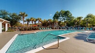 Located in Orange Beach, Alabama, this resort-style outdoor amenity area offers a generously sized swimming pool as its centerpiece, designed with a curvilinear shape and crystal-clear turquoise water. Decorative water fountain jets arc gracefully across the pool's surface, adding a lively, resort-quality aesthetic to the space. The expansive pool deck is finished in a light-toned concrete surface and is furnished with rows of bright teal lounge chairs positioned to take full advantage of the abundant Alabama sunshine. A dedicated grilling and outdoor dining area is equipped with full-sized gas grills, picnic tables, and shaded umbrella seating, making it ideal for outdoor gatherings. A covered patio area adjoins the main building — a warm terracotta-toned structure with white columns and large windows — providing a shaded retreat with additional lounge furniture. The entire area is framed by mature palm trees and towering pines that create a lush, tropical atmosphere and a sense of privacy. The overall design balances functionality with a welcoming, vacation-inspired ambiance, offering residents and guests a well-appointed outdoor living experience in the heart of the Gulf Coast.