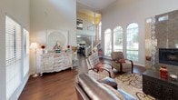 This single-floor clubhouse offers an impressive and inviting gathering space defined by its dramatic architectural proportions and thoughtful design. The main living area immediately draws the eye upward to soaring ceilings adorned with clerestory windows that flood the room with natural light from above, complementing the generously sized arched windows lining the walls and offering views of the surrounding greenery outdoors.

Rich, dark hardwood flooring flows throughout the space, providing a warm and polished foundation that anchors the room's elegant aesthetic. A standout feature is the floor-to-ceiling tile-surround fireplace, which serves as a striking focal point and lends a sense of comfort and sophistication to the seating area. The interior staircase visible from the main room adds architectural interest and reinforces the grand scale of the space.

The clubhouse is furnished with comfortable seating arrangements, including a leather sofa, patterned accent chairs, and a substantial dark wood trunk-style coffee table, all set atop a decorative area rug. Decorative consoles and sideboards with ornate detailing add character throughout. Multiple French door entries and large windows ensure seamless transitions between the interior and exterior, while the open layout and lofty ceilings create an airy, community-oriented atmosphere throughout.