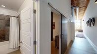 Located at 25 Draper St in Greenville, SC, this 2-bedroom, 2-bathroom loft showcases a striking industrial-chic aesthetic rooted in converted warehouse architecture. The open-concept living and kitchen area is anchored by soaring ceilings with exposed wooden beams and ductwork, polished concrete floors, and dramatic floor-to-ceiling factory windows that flood the space with natural light and frame lush outdoor views. Original exposed brick walls add warmth and character throughout.

The kitchen is well-appointed with rich espresso cabinetry, a subway tile backsplash, stainless steel appliances, and a granite-topped island with bar seating beneath a pair of industrial pendant lights. The living area offers an expansive, loft-style feel with generous room to entertain.

Each bedroom features soaring ceilings with transom-style clerestory windows, walk-in closets, and a serene ambiance. The spacious primary bedroom includes direct access to a dedicated closet. Both bathrooms are finished with dark slate and neutral tile surrounds, tub-shower combinations, and warm wood vanities with stone countertops.

A dedicated laundry room with full-size washer and dryer adds convenience. Stylish barn-style sliding doors and clean white walls complete the sophisticated yet urban atmosphere of this one-of-a-kind loft.