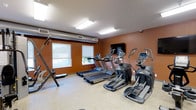 This fitness center is a dedicated exercise space designed with functionality and practicality in mind. The room features warm, earthy-toned walls that create an energizing atmosphere, complemented by light-toned flooring that is durable and easy to maintain. Overhead, flush-mounted fluorescent lighting ensures the space is brightly and evenly illuminated throughout the day. Two windows along the exterior wall allow natural light to filter in, adding to the open feel of the room. The space is outfitted with a lineup of modern cardio equipment, including multiple treadmills and elliptical cross-trainers from reputable fitness brands, as well as a multi-functional cable and weight machine positioned along one wall. A wall-mounted flat-screen display is centrally placed, allowing users to stay entertained during their workouts. Ventilation is well-addressed with visible ductwork and air vents integrated into the upper portion of the walls, ensuring proper airflow throughout the room. The layout is thoughtfully arranged to provide ample space between machines, allowing for comfortable and safe use. Overall, this fitness center offers a clean, focused environment ideal for cardiovascular training and general exercise.