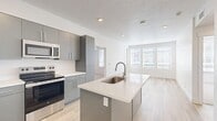 This well-designed 2-bedroom, 2-bathroom residence in Salt Lake City, Utah offers a thoughtful single-floor layout that maximizes functionality and comfort. The open-concept kitchen and living area serves as the heart of the home, featuring sleek gray flat-front cabinetry, white quartz countertops, a center island with a built-in sink, and a full suite of stainless steel appliances including a French door refrigerator, electric range, and microwave. Light wood-tone luxury vinyl plank flooring runs throughout the main living areas, complemented by recessed lighting and large windows dressed with horizontal blinds that fill the space with natural light. The living area offers additional windows with city views, enhancing the bright and airy ambiance. Both bedrooms are carpeted and well-proportioned, with the primary bedroom featuring a generous walk-in closet with built-in shelving and hanging rods. Each bathroom is finished with wood-look LVP flooring, a slate-blue vanity with white countertop, and chrome fixtures. A hallway connects the private areas of the home, maintaining a clean and organized flow. The overall aesthetic is clean, contemporary, and move-in ready.