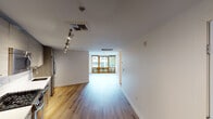 This single-floor unit offers a clean, contemporary design throughout, with warm hardwood flooring that flows seamlessly from room to room. The open living area is bright and airy, anchored by a striking set of floor-to-ceiling windows framed in rich wood tones that fill the space with abundant natural light and provide views of the surrounding streetscape and greenery. Track lighting overhead adds a functional, modern touch to the main living zone.

The kitchen is well-appointed with a full suite of stainless steel appliances, including a gas range, built-in microwave, dishwasher, and a large refrigerator. Flat-front cabinetry in a warm gray-brown finish lines the walls, complemented by a white tile backsplash and clean countertops, offering generous storage and prep space.

A secondary room features its own wood-framed window with views of mature trees, providing a quiet, naturally lit retreat. The bathroom is elegantly finished with warm-toned tile flooring, a double-sink vanity with an extended backlit mirror, ample under-counter storage, and a glass-enclosed walk-in shower with pebble stone flooring and full tile surround. The overall ambiance of the space is modern and refined, with a neutral palette that creates a bright, welcoming atmosphere throughout.