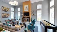 This single-floor clubhouse is thoughtfully designed to provide a welcoming and versatile gathering space for residents. The interior is anchored by a dramatic floor-to-ceiling orange accent wall that frames a sleek fireplace topped with a large flat-screen television, creating a bold focal point in the main lounge area. Soaring ceilings amplify the sense of openness, while an abundance of windows floods the space with natural light and offers views of the outdoor pool area.

The main lounge is furnished with plush sofas, teal wingback chairs, and stylish coffee tables, all set against soft grey walls and herringbone-patterned flooring. Decorative wall shelving adds a curated, gallery-like touch to the room. Adjacent seating areas feature additional armchairs and dining tables ideal for casual socializing.

A separate multi-purpose area showcases light wood-look tile flooring, high ceilings with a vibrant teal accent wall, modern bar-height bistro tables with teal stools, and framed artwork — lending an upbeat, energetic atmosphere perfect for social events or casual mingling.

The game room is equipped with a full shuffleboard table and is bathed in natural light through oversized windows, with views of the outdoor amenity area. Throughout the clubhouse, a cohesive palette of grey, teal, and warm amber tones ties the spaces together in a modern and inviting aesthetic.