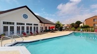 This single-floor community facility is centered around a pristine outdoor swimming pool, offering a resort-like atmosphere ideal for relaxation and recreation. The generously sized pool features crystal-clear turquoise water and is surrounded by a broad concrete sundeck furnished with lounge chairs and shaded seating areas complete with vibrant red umbrellas, perfect for unwinding on warm days.

The adjacent clubhouse building is a standout architectural feature, clad in classic white siding and topped with a steeply pitched dark roof. A distinctive circular window accents the upper gable facade, adding a charming architectural detail. Large, multi-pane French doors line the front of the building, flooding the interior with natural light and providing seamless access between the indoor and outdoor spaces.

The outdoor area is thoughtfully appointed with additional seating arrangements, including cushioned chairs grouped around tables for social gatherings. Mature trees in the background provide a natural backdrop and offer additional shade. The entire pool area is enclosed by a dark metal fence, ensuring both safety and privacy. The overall ambiance is bright, open, and inviting, combining classic architectural design with well-maintained outdoor amenities suited for community enjoyment throughout the warmer months.