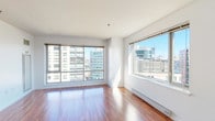 This well-appointed 1-bedroom, 1-bathroom unit is situated on an upper floor, offering sweeping urban skyline views through its expansive windows. The open layout is anchored by warm hardwood flooring that runs throughout the living areas and bedroom, reflecting abundant natural light and lending the space a bright, welcoming atmosphere.

The generous living area features large floor-to-ceiling windows on two walls, framing dynamic city views and flooding the interior with daylight. A baseboard heating system runs along the perimeter, ensuring comfort year-round. The space flows naturally toward the kitchen, which is separated by a breakfast bar counter. The kitchen is outfitted with rich wood cabinetry, white quartz countertops, a marble-style tile backsplash, stainless steel appliances including a dishwasher and range, and a window overlooking the cityscape.

The bedroom is a well-proportioned, quiet retreat with its own window providing additional natural light and urban views. A closet offers practical storage. In-unit laundry is conveniently tucked within the layout.

The bathroom features clean, white tile walls, a tub-shower combination, a wood-accented vanity with a mirror, and tile flooring, maintaining the modern aesthetic found throughout the unit.