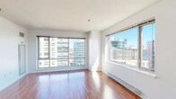 This well-appointed 1-bedroom, 1-bathroom unit is situated on an upper floor, offering sweeping urban skyline views through its expansive windows. The open layout is anchored by warm hardwood flooring that runs throughout the living areas and bedroom, reflecting abundant natural light and lending the space a bright, welcoming atmosphere.

The generous living area features large floor-to-ceiling windows on two walls, framing dynamic city views and flooding the interior with daylight. A baseboard heating system runs along the perimeter, ensuring comfort year-round. The space flows naturally toward the kitchen, which is separated by a breakfast bar counter. The kitchen is outfitted with rich wood cabinetry, white quartz countertops, a marble-style tile backsplash, stainless steel appliances including a dishwasher and range, and a window overlooking the cityscape.

The bedroom is a well-proportioned, quiet retreat with its own window providing additional natural light and urban views. A closet offers practical storage. In-unit laundry is conveniently tucked within the layout.

The bathroom features clean, white tile walls, a tub-shower combination, a wood-accented vanity with a mirror, and tile flooring, maintaining the modern aesthetic found throughout the unit.