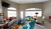 This single-floor space makes a strong impression through its generous ceiling height and well-proportioned layout. The living area is anchored by a bold, multicolored geometric area rug and furnished with a mix of teal accent chairs, a gray sofa with warm orange throw pillows, and a round dark wood coffee table — creating a lively yet cohesive aesthetic. The walls are finished in a neutral warm gray tone, providing a balanced backdrop for the colorful furnishings throughout.

Natural light is a defining feature of the space, delivered through large windows fitted with white horizontal blinds and accented by distinctive arched transom windows positioned high on the walls. These architectural details lend the room a sense of openness and volume. A glass-paneled entry door further enhances the flow of light.

Adjacent to the seating area, a compact dining space includes a round table paired with upholstered chairs in neutral tones, accented by a floral arrangement. The light wood-style flooring runs throughout, contributing warmth and continuity. Recessed ceiling lighting supplements the natural light, ensuring the space remains bright and welcoming at all times.