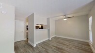 This single-floor unit offers a clean, modern aesthetic defined by consistent wood-look luxury vinyl plank flooring and neutral warm-gray walls throughout. The open-concept living area is the heart of the space, featuring a ceiling fan with integrated lighting and crisp white baseboards that frame the room neatly. A textured popcorn ceiling adds a subtle visual layer overhead.

The kitchen is partially separated from the living area by a half-wall partition that incorporates a pass-through window, providing both visual connectivity and functional convenience. Inside, the kitchen is outfitted with gray shaker-style cabinetry accented with gold-tone hardware, white countertops, a stainless steel sink, a tile backsplash, and a black refrigerator. The galley-style layout makes efficient use of the available space.

A spacious secondary room, suitable as a bedroom, provides ample wall space and includes a closet with a clean white door. The bathroom presents a simple, functional design with light wood-look tile flooring, a gray vanity with storage, a white toilet, and a bathtub with a curtain rod. Overall, the unit delivers a fresh, well-maintained environment with cohesive finishes and a practical layout.