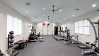Located at 511 Cobblestone Dr in Wilmington, NC, this well-appointed fitness center offers a clean, modern environment designed to support a wide range of workout routines. The single-floor facility features vaulted ceilings with recessed lighting and dual ceiling fans, creating a bright and airy atmosphere that keeps the space comfortable during exercise. Large windows along the walls allow natural light to complement the overhead lighting, contributing to an open and energizing ambiance. The flooring throughout consists of durable rubber gym matting, providing cushioning and traction for a variety of activities. The cardio zone is well-equipped with multiple treadmills and elliptical cross-trainers, offering users ample options for aerobic exercise. A dedicated functional training wall features TRX suspension straps and wall-mounted medicine balls, catering to bodyweight and resistance-based workouts. Additional strength and conditioning equipment rounds out the facility. The neutral white walls and streamlined layout keep the space feeling uncluttered and easy to navigate. Overall, this fitness center is thoughtfully designed to deliver a comprehensive workout experience in a clean, organized, and welcoming setting.