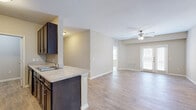 This single-story residence offers a practical and cohesive floor plan across three bedrooms, two bathrooms, a dedicated kitchen, and a combined kitchen/living area. Light wood-style flooring runs consistently throughout the main living spaces and bedrooms, contributing to a clean, unified aesthetic, while neutral beige tones on the walls create a warm, understated ambiance.

The kitchen is equipped with dark espresso cabinetry, stainless steel and black appliances, and a peninsula with a built-in sink and light-colored countertops. A fold-down breakfast bar adds a compact dining option. The adjoining living area benefits from a ceiling fan and direct access to the exterior through a glass door fitted with blinds, allowing natural light to fill the space.

A central hallway connects the three bedrooms, each featuring wood-style flooring, neutral walls, and individual closets with shelving. The primary bedroom includes a ceiling fan, an exterior-facing glass door, and an en-suite bathroom access point. Both bathrooms are finished with large-format tile flooring, dark vanity cabinetry, light countertops, and well-lit mirrors. The layout is further supported by three closets distributed throughout the unit, enhancing overall storage capacity.