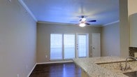 This single-story Dallas property offers a well-appointed open-concept living area that blends functional design with clean, modern finishes. The main living space is characterized by dark hardwood-style flooring that contrasts attractively against neutral-toned walls, creating a warm yet contemporary atmosphere. Crown molding runs along the ceiling perimeter, adding a refined architectural detail throughout the room.

A ceiling fan with an integrated light fixture is centrally positioned, providing both air circulation and ambient lighting to the space. Multiple windows fitted with horizontal blinds line one wall, allowing natural light to filter in while maintaining privacy. A glass-panel entry door is situated between the windows, contributing additional light to the interior.

The living area flows naturally toward the kitchen, where a granite countertop is visible at the edge of the frame, featuring a speckled pattern in neutral tones that complements the overall color palette. A secondary interior door provides additional access within the space. The single-floor layout ensures easy accessibility throughout, making this Dallas property a practical and attractive option.