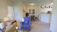 This single-floor residence offers a welcoming and efficiently designed layout that seamlessly connects the main living areas. The living room is carpeted in a soft, neutral tone and is furnished with comfortable seating, creating a warm and inviting atmosphere. Framed artwork adorns the walls, adding a personal touch to the space. Adjacent to the living area, a dedicated dining space accommodates a wrought-iron table and chairs, providing an intimate setting for meals. Decorative framed prints and a lush indoor plant enhance the corner, lending a touch of greenery to the interior. A half-wall pass-through connects the dining area to the kitchen, which features white cabinetry and is accented by a charming "Bistro" sign, giving the space a casual, café-inspired character. A paneled door leads to additional areas of the residence, maintaining a clean and classic aesthetic throughout. The overall ambiance is calm and residential, with consistent warm wall tones, soft lighting, and thoughtful décor that contribute to a comfortable and livable environment.