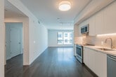 This thoughtfully designed single-floor unit offers a clean, contemporary aesthetic defined by crisp white walls, smooth ceilings, and rich dark hardwood flooring that flows seamlessly throughout the main living areas. The open-concept layout connects the kitchen and living space, creating an expansive, airy feel enhanced by natural light streaming through large, framed windows that offer views of the surrounding urban landscape.

The kitchen is a standout feature, showcasing floor-to-ceiling flat-front white cabinetry, a herringbone-patterned tile backsplash with under-cabinet lighting, and a full suite of stainless steel appliances including a range, microwave, dishwasher, and refrigerator. A single-basin sink with a modern faucet and white quartz countertops complete the polished look.

The living area is generously proportioned, with track lighting overhead and clean wall surfaces that offer flexible arrangement possibilities. A hallway with white interior doors connects the main spaces to a private bedroom, which features a large window allowing ample natural light.

The bathroom is finished in a neutral palette, featuring large-format dark tile flooring, a white vanity with quartz countertop, and a full soaking tub and shower combination surrounded by light stone-toned tile. A recessed ceiling light adds to the bright, clean atmosphere. Throughout the unit, every detail reflects a commitment to modern, minimalist design.