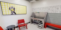 This 230-square-foot single-room pet grooming suite is thoughtfully designed with both functionality and whimsy in mind. The space features a classic white mosaic tile floor with black dot accents, providing a clean and durable surface suited for professional grooming use. A standout design element is the accent wall adorned with an oversized dog bone pattern wallcovering, complemented by a matching bone-shaped ceiling light fixture that adds a playful character to the room. The grooming area is equipped with a stainless steel elevated bathing tub complete with entry stairs, a wall-mounted dryer unit with a flexible hose, and a dark-surfaced grooming table, providing a fully operational workstation. Sage green vertical tiles line the lower half of the grooming station wall, topped by a charming illustrated wallpaper border featuring dog illustrations. The ceiling incorporates a gray and white checkerboard acoustic tile pattern. A small waiting nook near the entrance includes two red modern chairs, a round black side table, and a framed Dalmatian art print, creating a welcoming space for pet owners. A bold yellow ceiling accent above the entry area adds a vibrant finishing touch to this professionally equipped and creatively designed grooming suite.