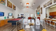 Located at 5500 Sampson St in Houston, TX, this single-floor community clubhouse offers a well-appointed and inviting space designed for socializing and relaxation. The living room is the centerpiece of the space, boasting dramatic vaulted ceilings that lend an open, airy feel. Large French doors with transom windows flood the area with natural light while providing views of the surrounding outdoor spaces. A tile-surround fireplace serves as a striking focal point, framed by decorative wall art and a mounted flat-screen television. The living area is thoughtfully furnished with a plush sofa, vibrant orange accent chairs, and a patterned area rug that adds warmth and color. A full-size billiards table provides an entertaining feature, complemented by built-in cabinetry and additional wall-mounted screens along one side of the room. Wood-look flooring runs throughout, enhancing the contemporary aesthetic. Pendant lighting in bold orange adds a pop of color overhead. The kitchen is equipped with stainless steel appliances, including a side-by-side refrigerator with an ice and water dispenser, a microwave, and warm wood cabinetry with ample counter space. Together, these spaces create a functional and stylish clubhouse environment ideal for community gatherings.