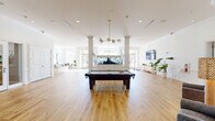 This expansive single-floor community amenity space offers a bright, open-concept layout finished with warm wide-plank hardwood flooring and crisp white walls throughout. The main living area is anchored by a striking built-in aquarium centerpiece flanked by white decorative columns, with a modern brass multi-globe chandelier suspended overhead. A full-size billiards table commands the center of this generous gathering space, making it an ideal hub for recreation and socializing. Elegant crown molding, recessed lighting, and large multi-pane French doors and windows flood the interior with natural light and offer views to the outdoors. A dedicated lounge nook features floor-to-ceiling windows with a bar-height counter and seating, perfect for casual work or conversation. A grand piano positioned near the windows adds a refined, cultural touch to the space. The built-in coffee and refreshment station, complete with illuminated glass cabinetry and a cushioned bench, provides a welcoming respite. A separate utility room houses smart package lockers and a refrigerated delivery unit for added convenience. Throughout, the design balances classic architectural details with modern furnishings, creating an inviting, multi-functional environment.
