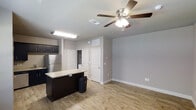 This single-floor unit in White Settlement offers a clean, modern design with a practical and open layout. The living and kitchen areas flow seamlessly together, creating a cohesive and functional space. The kitchen is outfitted with dark cabinetry that provides a striking contrast against the light-toned walls, and a center island with a light stone countertop serves as both a prep area and a casual gathering spot. A stainless steel refrigerator adds a contemporary touch to the kitchen's overall aesthetic. Durable wood-look plank flooring runs throughout the main living area, offering both style and easy maintenance. The living space is bright and airy, with neutral gray walls that provide a versatile backdrop for a variety of décor styles. A ceiling fan with an integrated light fixture keeps the space comfortable year-round. Multiple doors visible from the main living area suggest convenient access to additional rooms or storage spaces. Recessed and overhead lighting ensures the interior is well-illuminated throughout. The unit's straightforward, efficient design makes it a practical and attractive option for those seeking a well-appointed space in White Settlement.