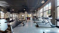 Located at 205 S High St in the heart of downtown Columbus, Ohio, this fitness facility offers a generously proportioned, single-floor layout that accommodates a full range of workout zones. The space is defined by its industrial-modern aesthetic, featuring exposed ductwork, polished concrete floors, and soaring ceilings that lend an open, energetic atmosphere throughout.

Floor-to-ceiling windows line multiple walls, flooding the interior with abundant natural light while providing views of the surrounding urban streetscape. A large glass garage door serves as a dramatic architectural feature in one zone, reinforcing the industrial character and connecting the interior to the outdoors.

The cardio area is equipped with a row of treadmills, stationary bikes, and cycling spin bikes positioned along the window-facing wall. A dedicated free-weight and functional training zone houses kettlebells, medicine balls, dumbbells, and benches, with a motivational "One Hour for the Other 23" mural as a striking focal point. Selectorized cable and strength machines occupy their own section, while an open rubber-floored training area provides space for dynamic movement and group workouts.

A living green wall installation adds a natural, biophilic element to the space, complemented by a front reception and check-in desk area. The facility is well-lit with pendant and track lighting throughout, creating a polished yet functional environment designed to support comprehensive fitness programming.
