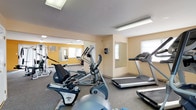 This fitness center offers a comprehensive range of exercise equipment suited for both cardio and strength training. The space is organized into distinct workout zones, with warm yellow accent walls and neutral carpeted flooring throughout. Overhead fluorescent lighting keeps the area well-lit, while multiple windows allow natural light to filter in, creating a bright and energizing atmosphere.

The cardio section is equipped with treadmills, elliptical trainers, and a stationary recumbent bike, providing a variety of low- and high-impact exercise options. A multi-station cable weight machine, accompanied by an adjustable workout bench, anchors the strength training area and supports a wide range of resistance exercises. A dedicated free weights zone features a tiered dumbbell rack with an assortment of weights and an additional flat bench for supplementary exercises.

Large wall-mounted mirrors are strategically placed throughout the space, enhancing the sense of openness and allowing users to monitor their form during workouts. A wall-mounted air conditioning unit ensures comfortable temperatures year-round. The layout is thoughtfully arranged to allow ample circulation between equipment, making the fitness center functional and accessible for a variety of training routines.