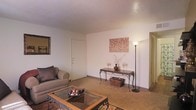 This single-floor space features a living area finished in warm, neutral tones that create an inviting and relaxed atmosphere. The room is characterized by light beige carpet flooring and smooth white walls accented by a textured ceiling, contributing to a cozy residential feel. Natural light is complemented by a tall floor lamp, ensuring the space remains well-lit throughout. The living area comfortably accommodates a cushioned sofa paired with a glass-top coffee table supported by ornate wrought-iron legs, adding a decorative touch to the space. Against the far wall, an elegant wrought-iron console table serves as a focal point, styled with decorative vases, candle holders, and a woven storage basket beneath. A framed wall art piece featuring a botanical motif adds a subtle artistic element. A framed mirror on the adjacent wall enhances the sense of space. Doorways on either side of the room suggest additional areas beyond the main living space, offering functional connectivity throughout the unit. Overall, the space presents a warm, understated aesthetic with thoughtful decorative details.