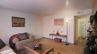 This single-floor space features a living area finished in warm, neutral tones that create an inviting and relaxed atmosphere. The room is characterized by light beige carpet flooring and smooth white walls accented by a textured ceiling, contributing to a cozy residential feel. Natural light is complemented by a tall floor lamp, ensuring the space remains well-lit throughout. The living area comfortably accommodates a cushioned sofa paired with a glass-top coffee table supported by ornate wrought-iron legs, adding a decorative touch to the space. Against the far wall, an elegant wrought-iron console table serves as a focal point, styled with decorative vases, candle holders, and a woven storage basket beneath. A framed wall art piece featuring a botanical motif adds a subtle artistic element. A framed mirror on the adjacent wall enhances the sense of space. Doorways on either side of the room suggest additional areas beyond the main living space, offering functional connectivity throughout the unit. Overall, the space presents a warm, understated aesthetic with thoughtful decorative details.